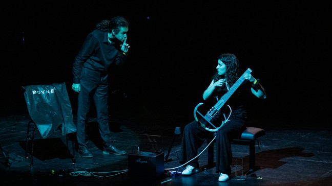 Eine Musikerin sitzt Gitarre spielend auf der Bühne und wird von ihrem Mit-Performer mit Handzeichen zum Leisesein ermahnt. Die Performance von Antigone Baxe behandelt das Thema Redefreiheit in unserer Gesellschaft. | Bildquelle: © Jakob Schad