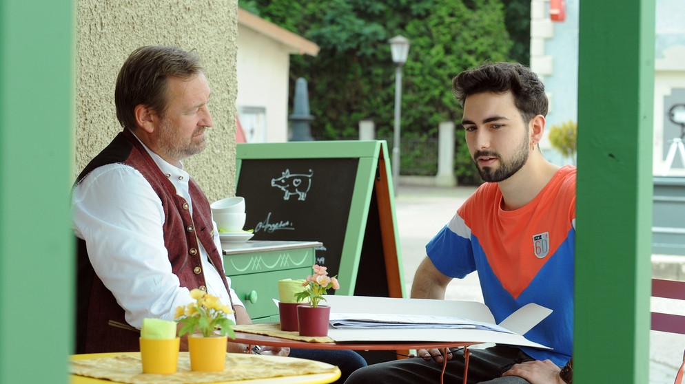 Dahom is Dahoam: Gregor (Holger Matthias Wilhelm, links) sitzt mit Felix (Shayan Hartmann, rechts) im Schanigarten. | Bildquelle: BR/Marco Orlando Pichler
