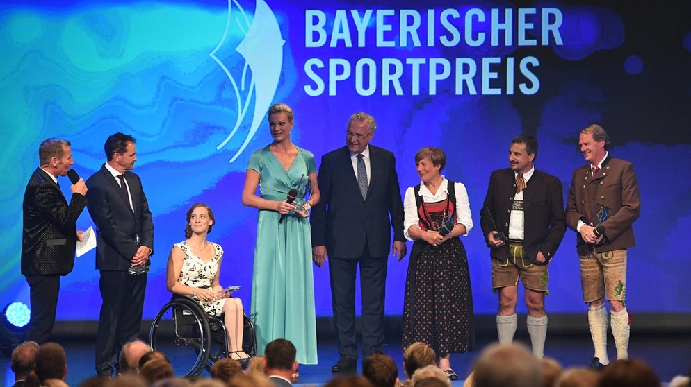 Verleihung des Bayerischen Sportpreises in der BMW Welt in München. Der Moderator Markus Othmer (l-r), Nordischer Kombinierer Hermann Weinbuch, Monoskibobfahrerin Anna Schaffelhuber, die ehemalige Skirennläuferin, Maria Höfl-Riesch, der bayerische Innenminister Joachim Herrmann (CSU), die ehemalige Skirennläuferin Rosi Mittermaier-Neureuther, der ehemalige Rodler Georg "Schorsch" Hackl und der ehemalige Skifahrer Markus Wasmeier stehen mit dem Bayerischen Sportpreis auf der Bühne. | Bildquelle: dpa-Bildfunk/Felix Hörhager 