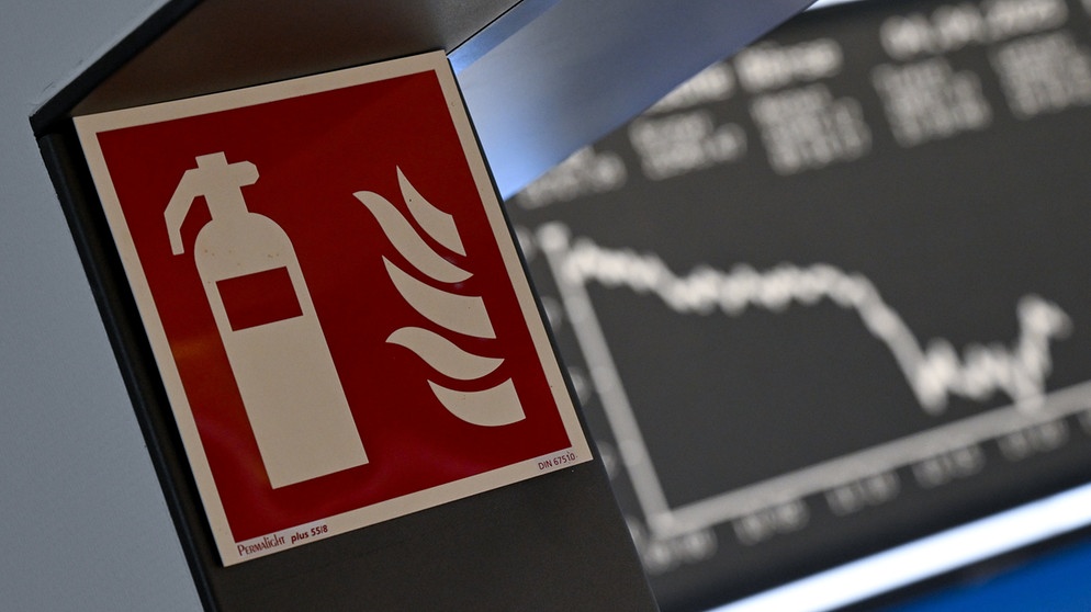 Ein Hinweisschild für einen Feuerlöscher ist an einem Durchgang der Frankfurter Wertpapierbörse angebracht, während die Anzeigetafel mit der Dax-Kurve fallende Kurse anzeigt. | Bildquelle: Arne Dedert/dpa-Bildfunk