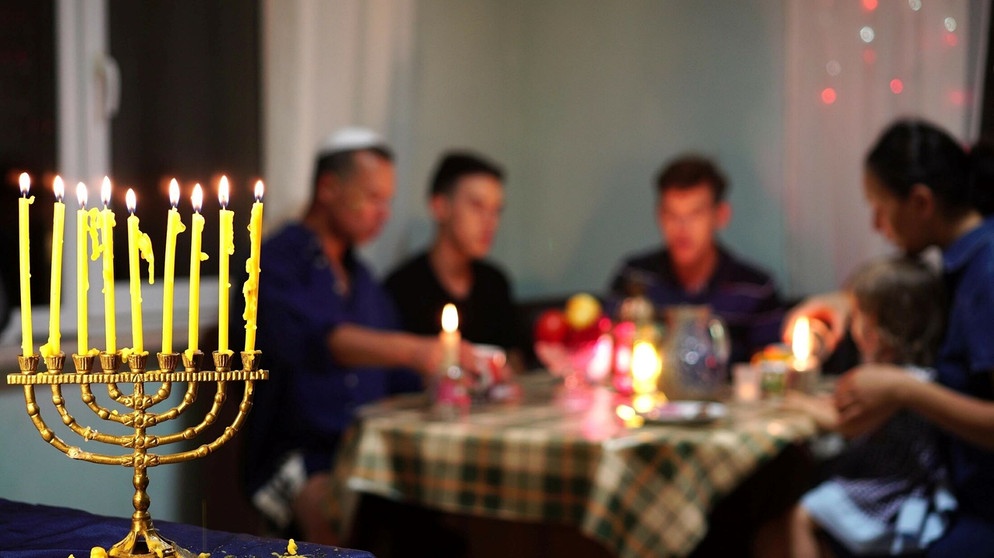 Ein "Chanukkaleuchter" bzw. Neunarmiger Kerzenständer in einer Wohnung. Im Hintergrund ist eine Familie zu Tisch. | Bildquelle: stock.adobe.com/Fevziie