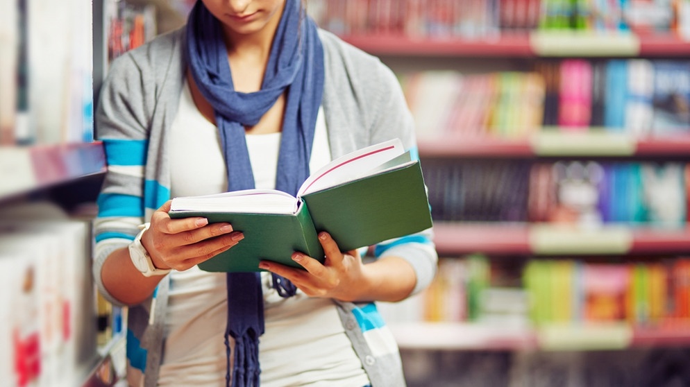 Eine Frau mit einem Buch in der Hand steht vor einem Bücherregal | Bildquelle: colourbox.com