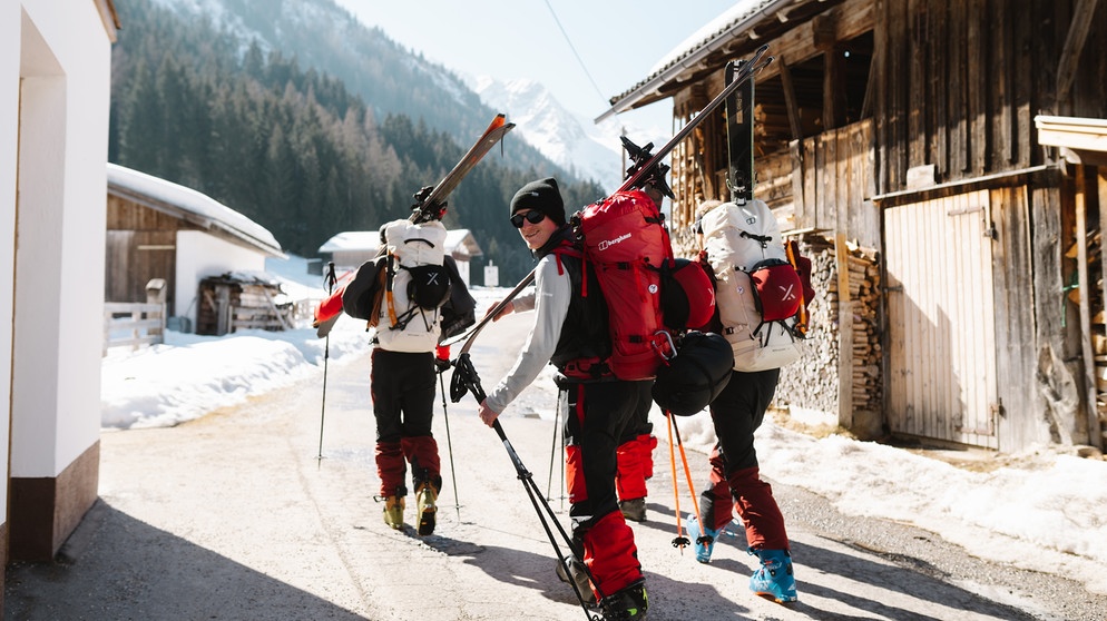Drei Skifahrer mit Gepäck in einem Dorf zu Fuß unterwegs  | Bildquelle: Moritz Ablinger