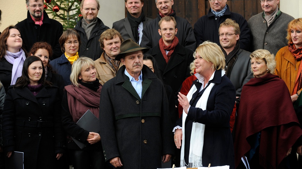 Bürgermeister Schattenhofer hofft, dass der Schwindel der Lansinger, mit dem sie sich den Auftritt des BR-Chors erschlichen haben, nicht auffliegt. Von links: Bürgermeister Schattenhofer (Werner Rom) mit der Organisatorin (Madeleine Vester) und dem Chor des BR. | Bildquelle: BR/Marco Orlando Pichler