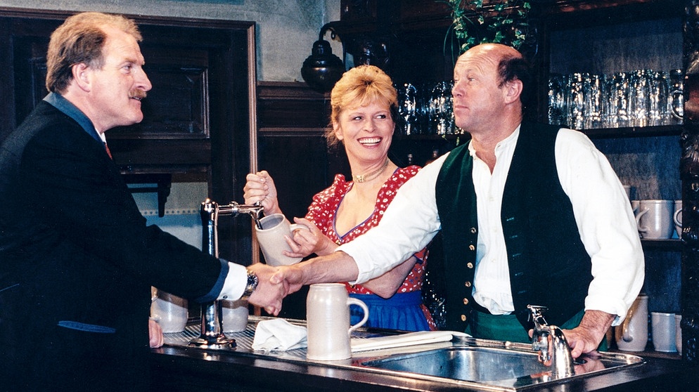 Chiemgauer Volkstheater - Der Witwentröster: Rosa (Mona Freiberg) und Simmerl (Hans Stadlbauer) stehen hinter der Bar. Simmerl schüttelt Antons (Bernd Helfrich) Hand. | Bildquelle: BR/Sessner