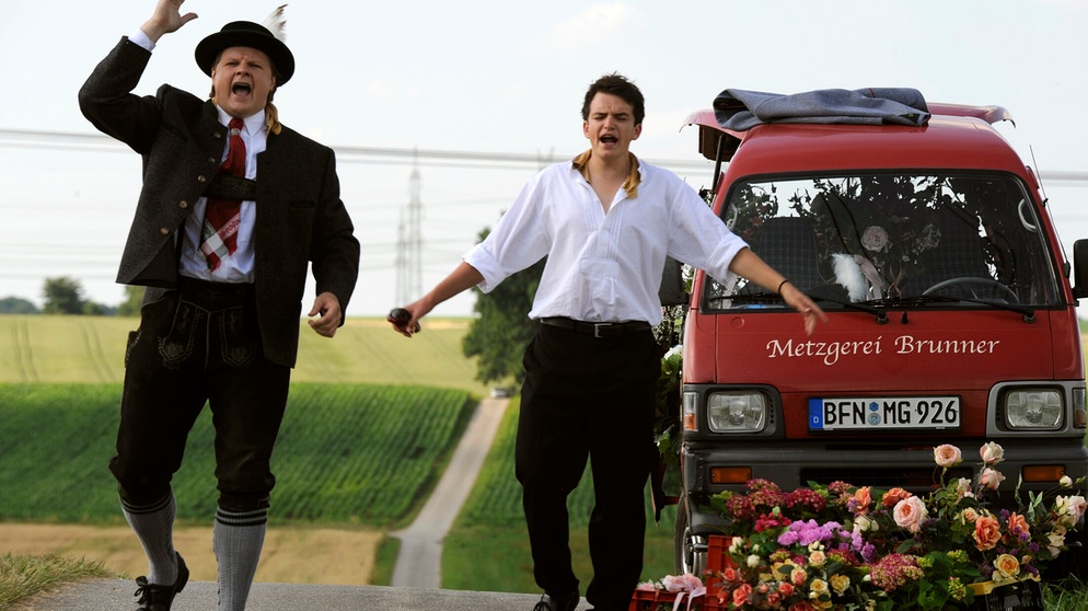 Dahoam is Dahoam. Keiner hält an, um Max und Florian mit dem liegen gebliebenen Auto zu helfen. Von links: Max (Michael A. Grimm) und Florian Brunner (Tommy Schwimmer). | Bildquelle: BR/Marco Orlando Pichler