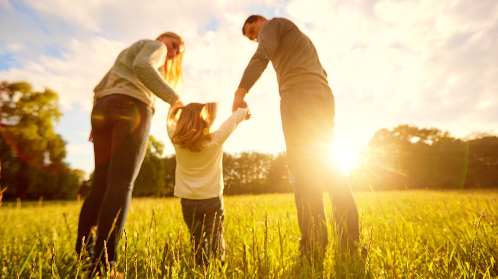 Zwei Eltern mit ihrer Tochter an der Hand auf einem Feld im Gegenlicht. | Bildquelle: stock.adobe.com/Studio Romantic