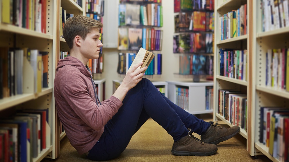 Ein junger Mann sitzt zwischen Bücherregalen auf dem Boden und liest ein Buch | Bildquelle: colourbox.com / Dmitrii Shironosov