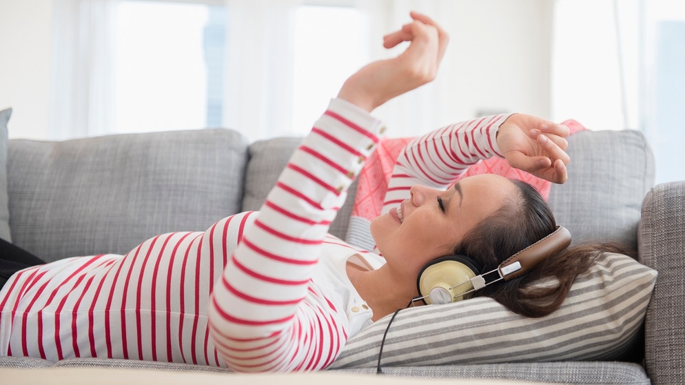 Eine junge Frau liegt auf dem Sofa. Sie hört über Kopfhörer Musik und bewegt die Arme dazu. | Bildquelle: mauritius-images