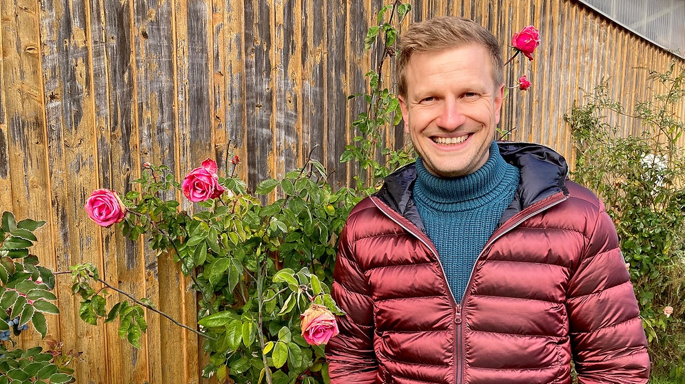 Moderator Florian Kienast vor einem Stadel, an dem eine Staudenrose blüht (aufgeommen am 08.12.2022) | Bildquelle: BR