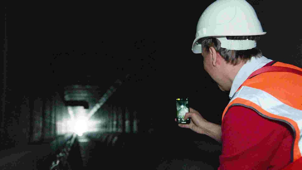 Mann in Schutzkleidung fotografiert mit seinem Handy Wärmetauschermodule in einem Abwasserkanal. Ansicht von hinten.
| Bildquelle: BR