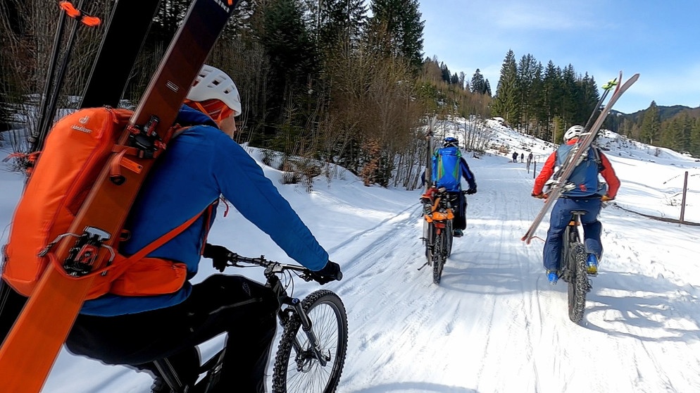 Drei Mountainbiker mit Skiern auf den Rucksäcken fahren durch ein verschneites Tal | Bildquelle: BR