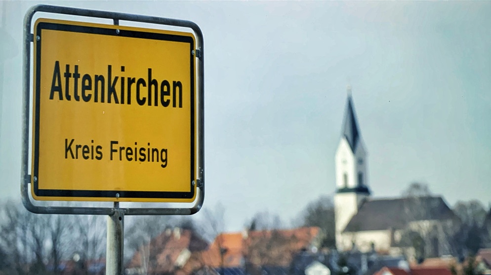 Ortsschild mit der Aufschrift "Attenkirchen"; im Hintergrund ist der Kirchturm von Attenkirchen zu sehen. Weil es nach einem Großbrand im Jahr 1762 keine Archivbestände mehr gab, galt das Hallertauer Dorf Attenkirchen lange Zeit als "Ort ohne Geschichte". | Bildquelle: BR/Michael Mandlik