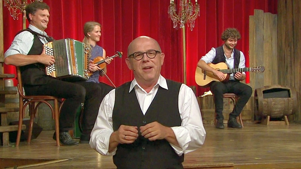 Gastgeber Jürgen Kirner bei der Volkssängerrevue Brettl-Spitzen XII. Im Hintergrund die Brettl-Spitzen-Musi auf der Bühne (Bernhard Gruber, Katharina Baur, Bernhard Filser). | Bildquelle: Bayerischer Rundfunk